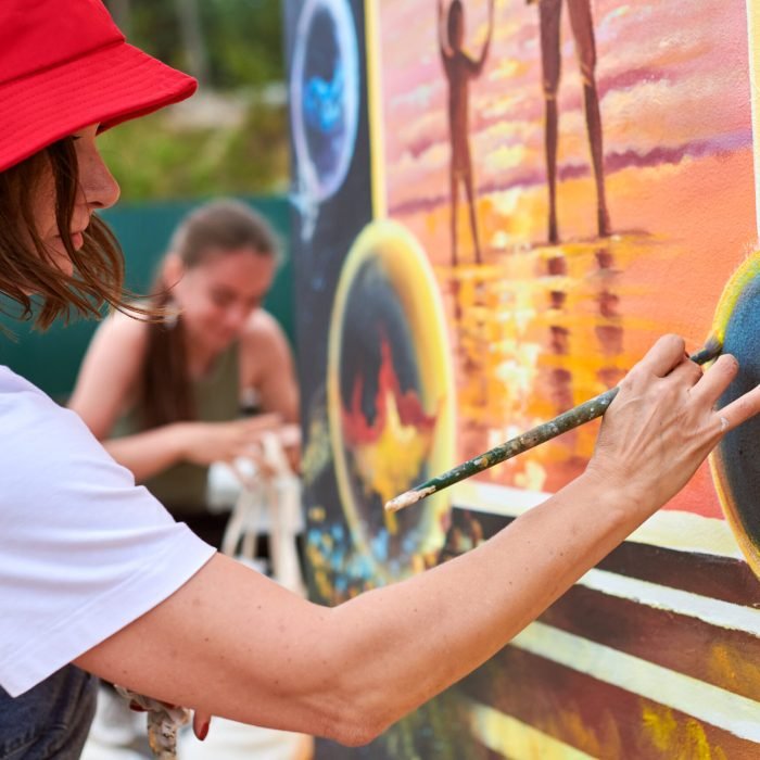 Female painter in red hat draws picture with paintbrush on canvas for outdoor street exhibition, close up side view of female artist apply brushstrokes to canvas, symphony of art creativity